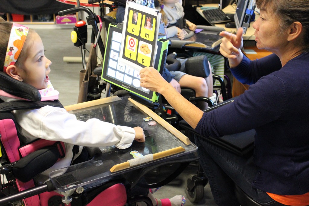 An image of a teacher holding a card up to a student. The card has images and the letters a, m, t, p on it, each letter corresponding to one of the images