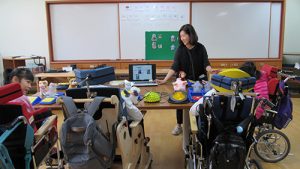 Image of a teacher standing in front of a group of four children in wheelchairs