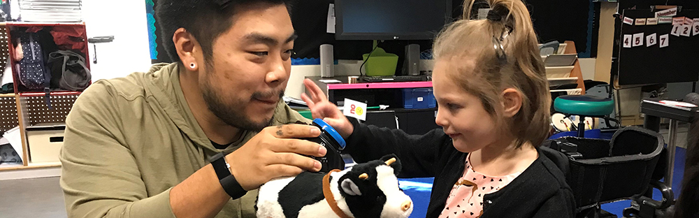 An image of a student pressing a large blue button being held up by a teacher