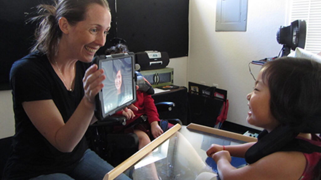 Image of a teacher holding up a tablet  displaying a smiling face on it, and showing it to a smiling student 