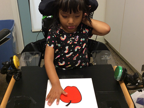 Image of a child in a wheelchair placing her hand on a picture of an apple lying on the table in front of her
