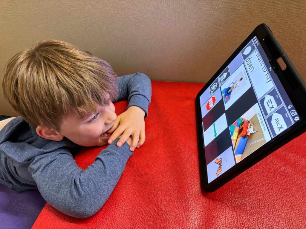 A young boy resting his arms and chin on the edge of a table, looking at images on a tablet in front of him