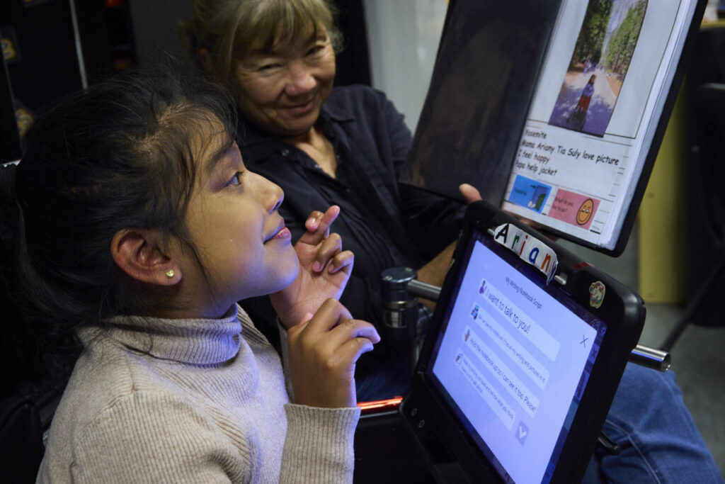 An instructor shows a student a page in a binder that has an photo with a question beneath it and two text and image cards that the student could point to in answer to the question