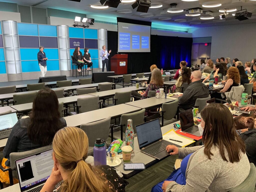 Image of a conference workshop in progress with four presenters on stage and several people sitting at long tables in the audience
